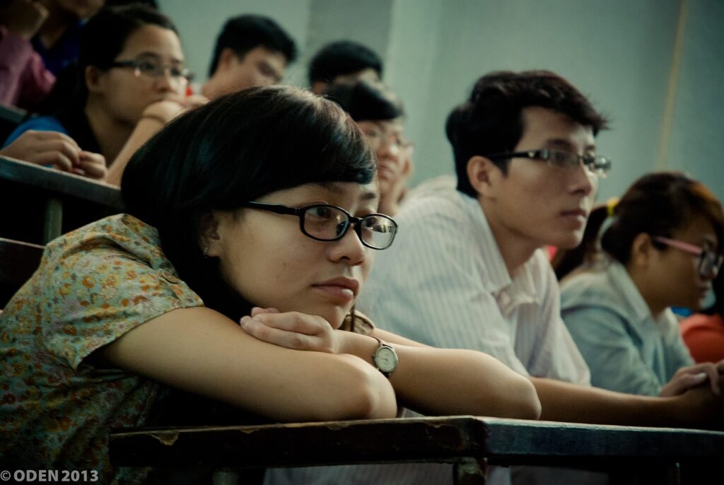 A young woman wearing glasses, resting her chin on her hands in a classroom, appearing deep in thought.