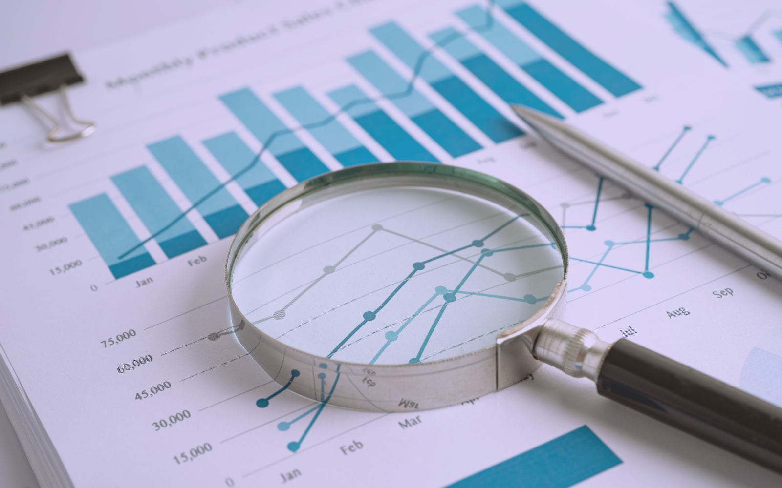 An image of "data analysis" featuring charts with bar and line graphs, a magnifying glass, pen, and paperclip on a report, emphasizing detailed data examination.