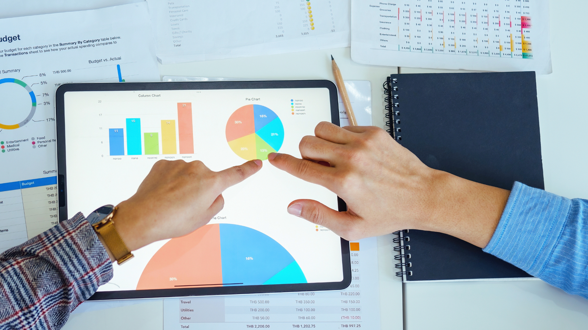 Two hands pointing at a tablet displaying budget charts, including column and pie charts, with papers and a notebook on a desk, analyzing spending categories.