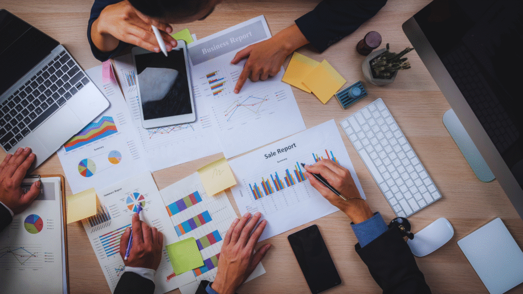 Business team analyzing charts and graphs on papers and a tablet, with laptops, sticky notes, and a keyboard on a wooden desk.