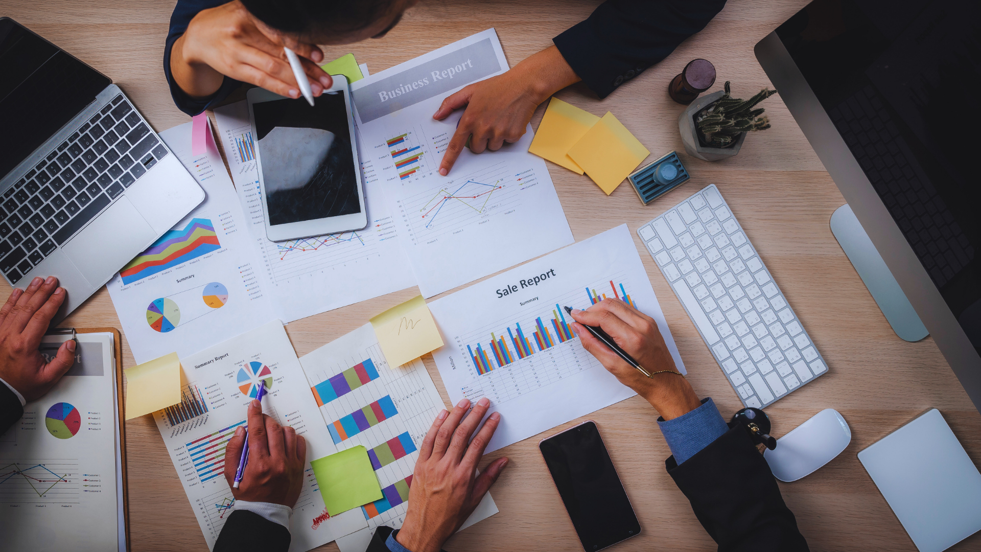 Business team analyzing charts and graphs on papers and a tablet, with laptops, sticky notes, and a keyboard on a wooden desk.