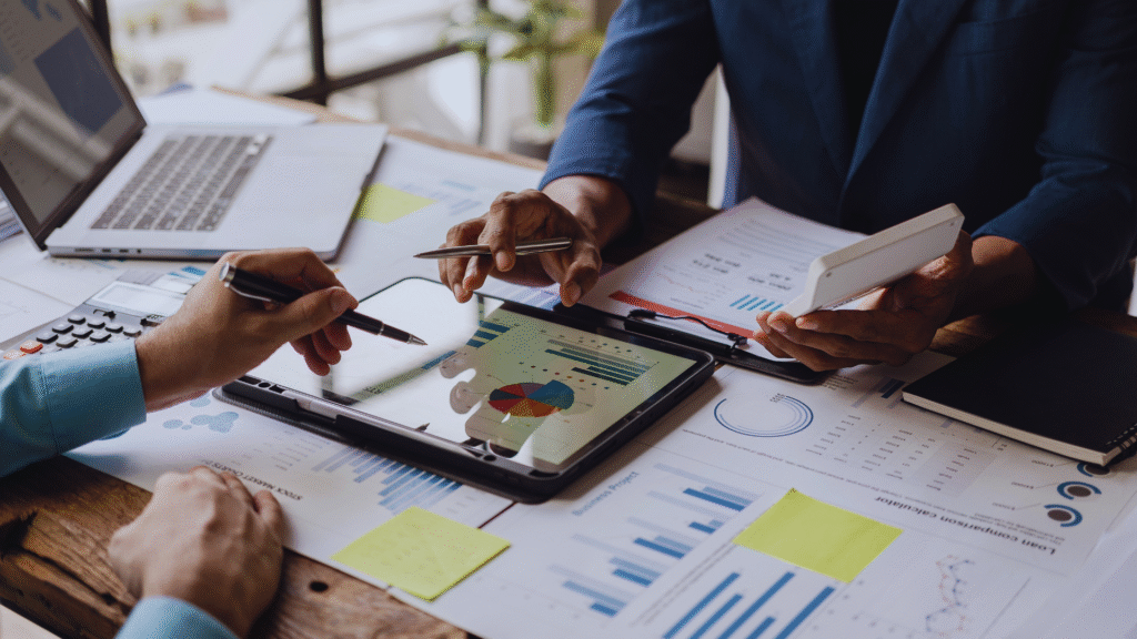Business meeting with professionals analyzing charts and graphs on a tablet, surrounded by laptops, papers, and calculators.