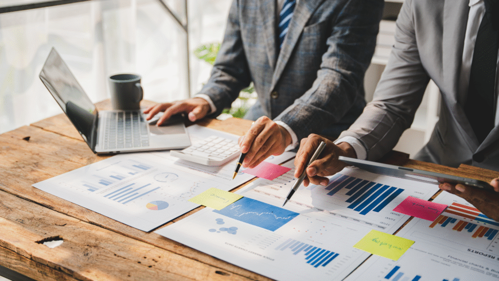 Two business professionals analyzing charts and graphs on a wooden table with a laptop, calculator, coffee mug, and sticky notes.