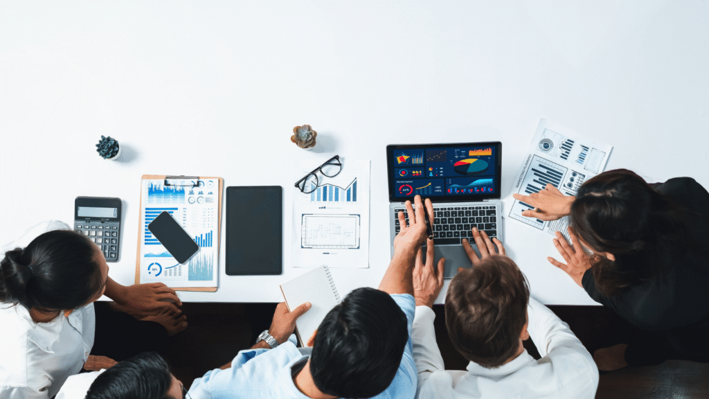 A team collaborating around a table with a laptop displaying charts, surrounded by papers, a calculator, and a clipboard with graphs.