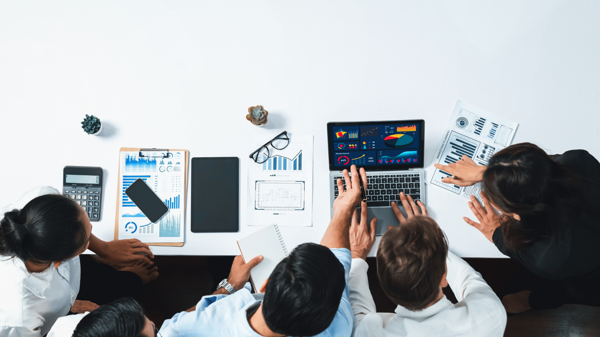 A team collaborating around a table with a laptop displaying charts, surrounded by papers, a calculator, and a clipboard with graphs.